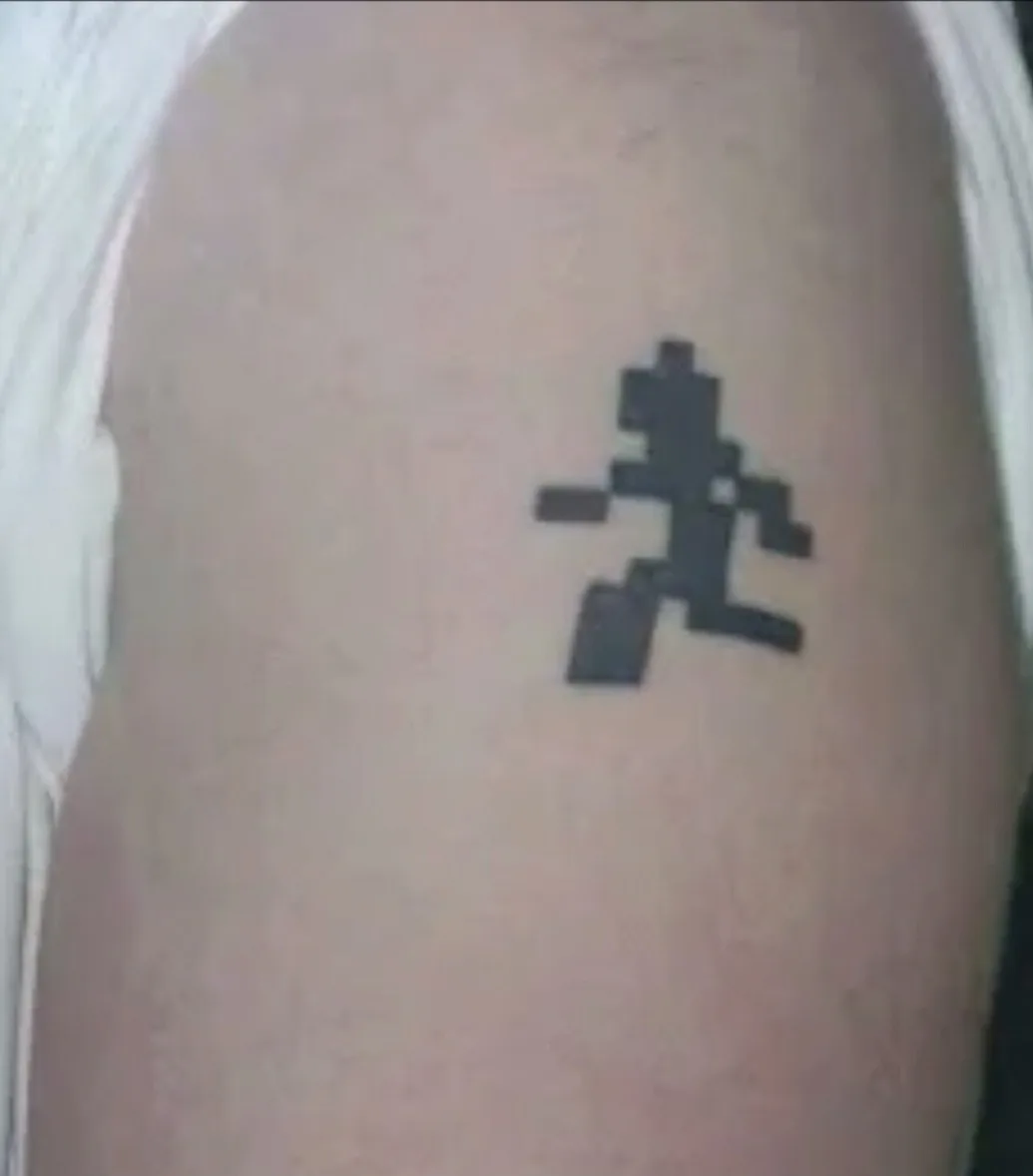 A close-up photo shows a small black pixel art tattoo on a person's upper arm, depicting a blocky, 8-bit style character in…