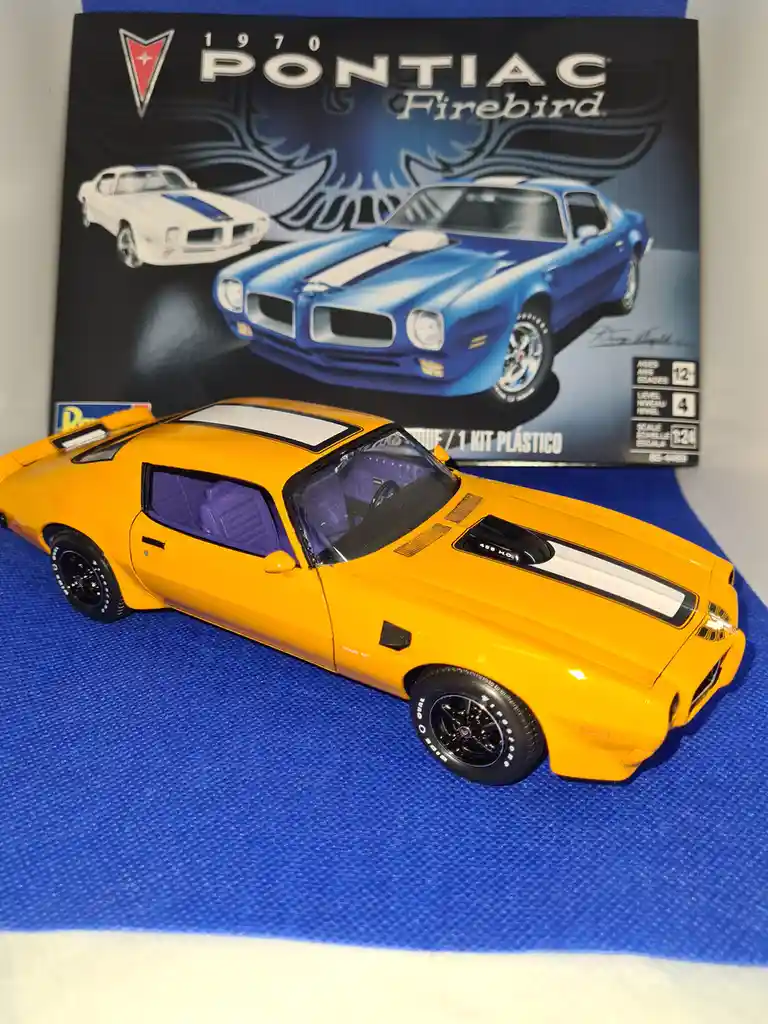 A bright yellow 1:24 scale plastic model of a 1970 Pontiac Firebird sits on a blue textured surface in front of its original…