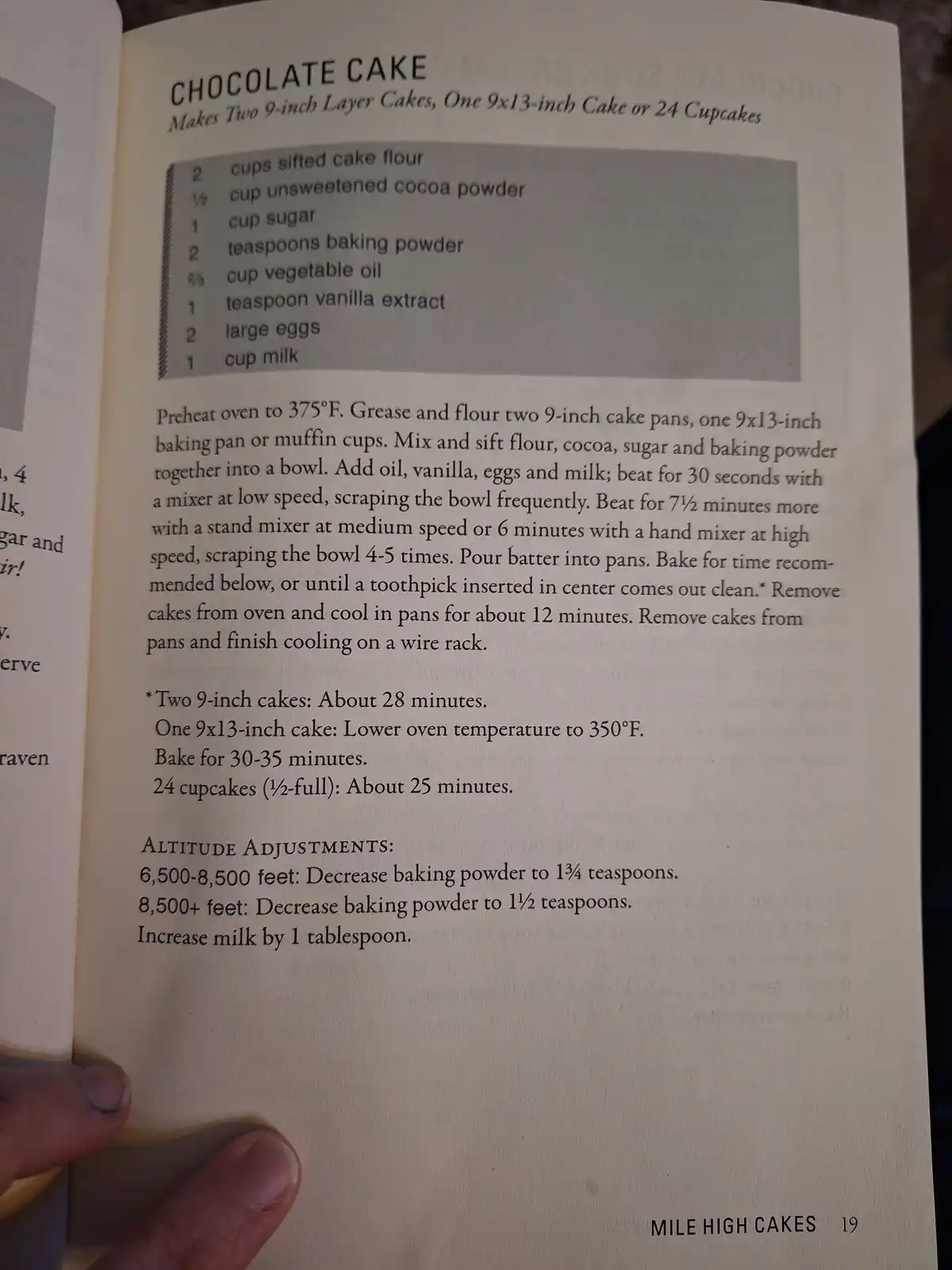 A close-up photo of a cookbook page titled 'CHOCOLATE CAKE' showing ingredients and baking instructions, with a hand holding…