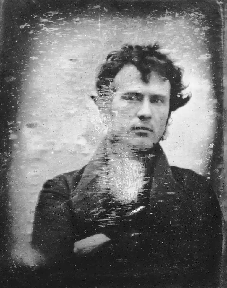 A grainy, vintage black-and-white portrait of a man with dark, curly hair and a serious expression, wearing a dark jacket…