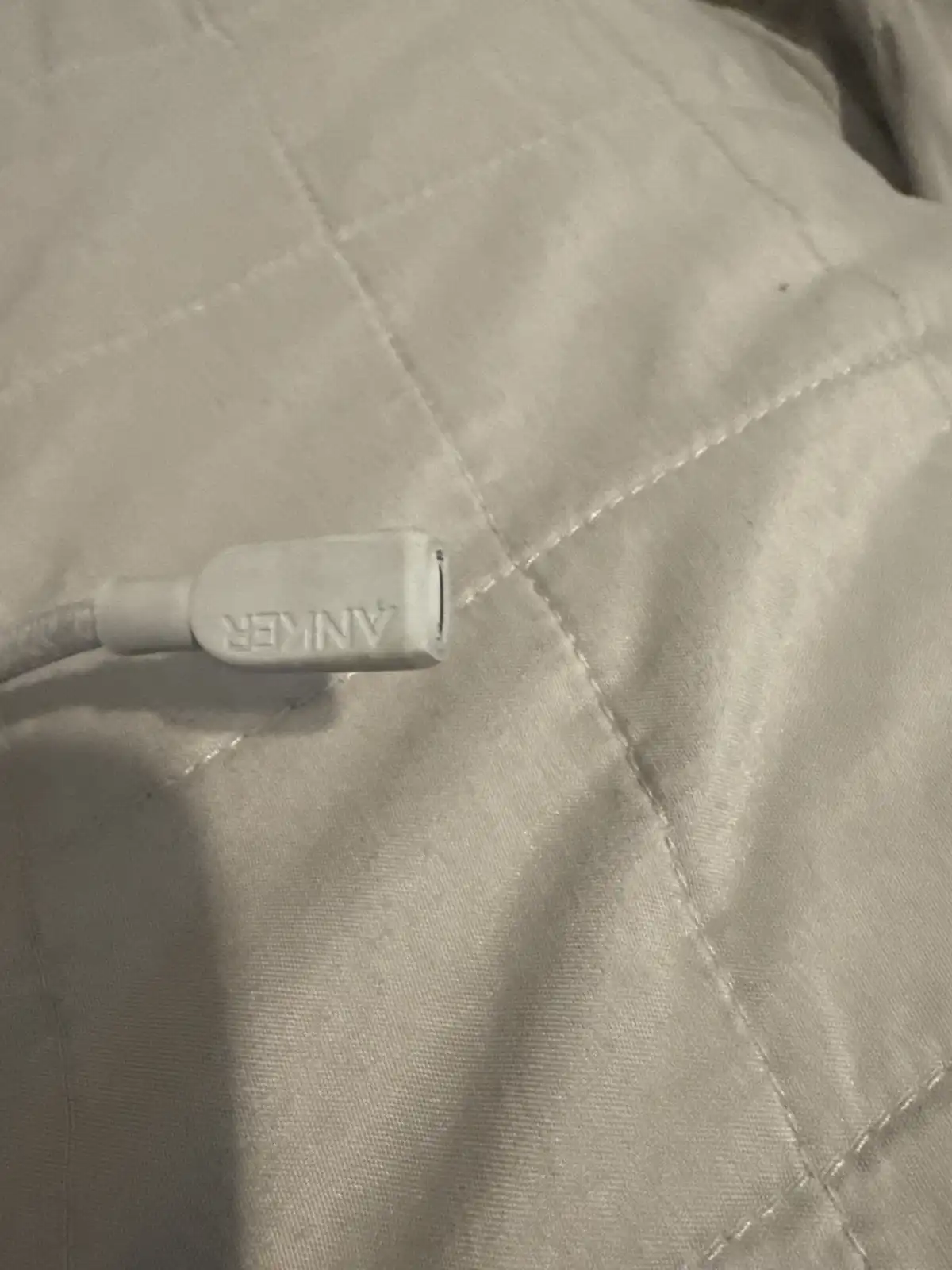 A close-up photo shows a white Anker USB cable plug resting on a light-colored, quilted fabric surface. The brand name…