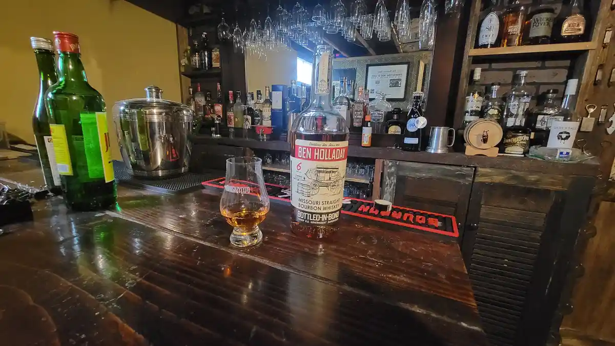 A close-up shot of a bar counter featuring a bottle of Ben Holladay Missouri Straight Bourbon Whiskey and a partially filled…