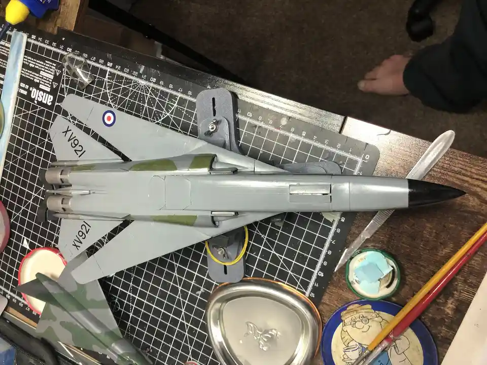 A partially painted model jet fighter, marked with 'XV921' and a roundel, sits on a black gridded cutting mat surrounded by…