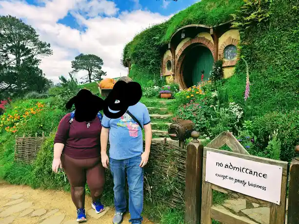 Two people stand in front of a hobbit-style house with a green roof and arched door, surrounded by lush gardens and flowers…