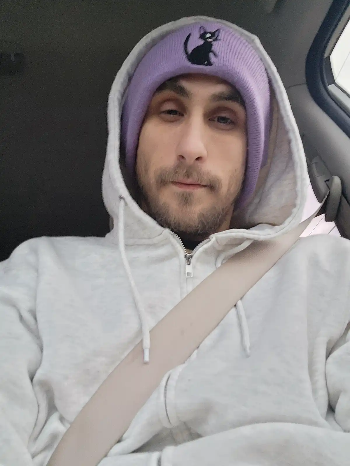 A man with a light beard sits in a car seat wearing a white zip-up hoodie with the hood up, and a purple beanie with a black…