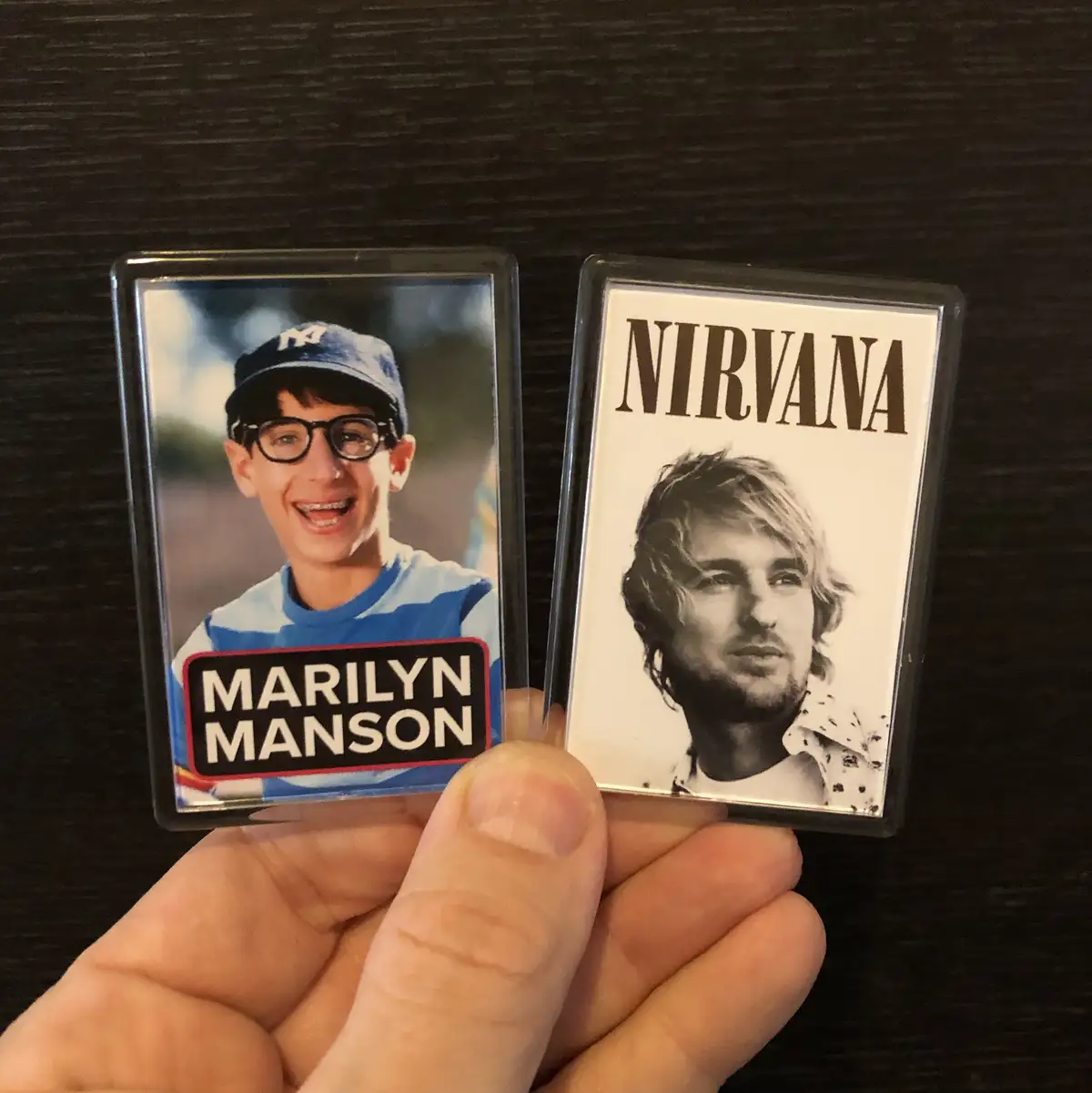 A hand holds two rectangular fridge magnets against a dark wood surface. The left magnet features a photo of a young boy…