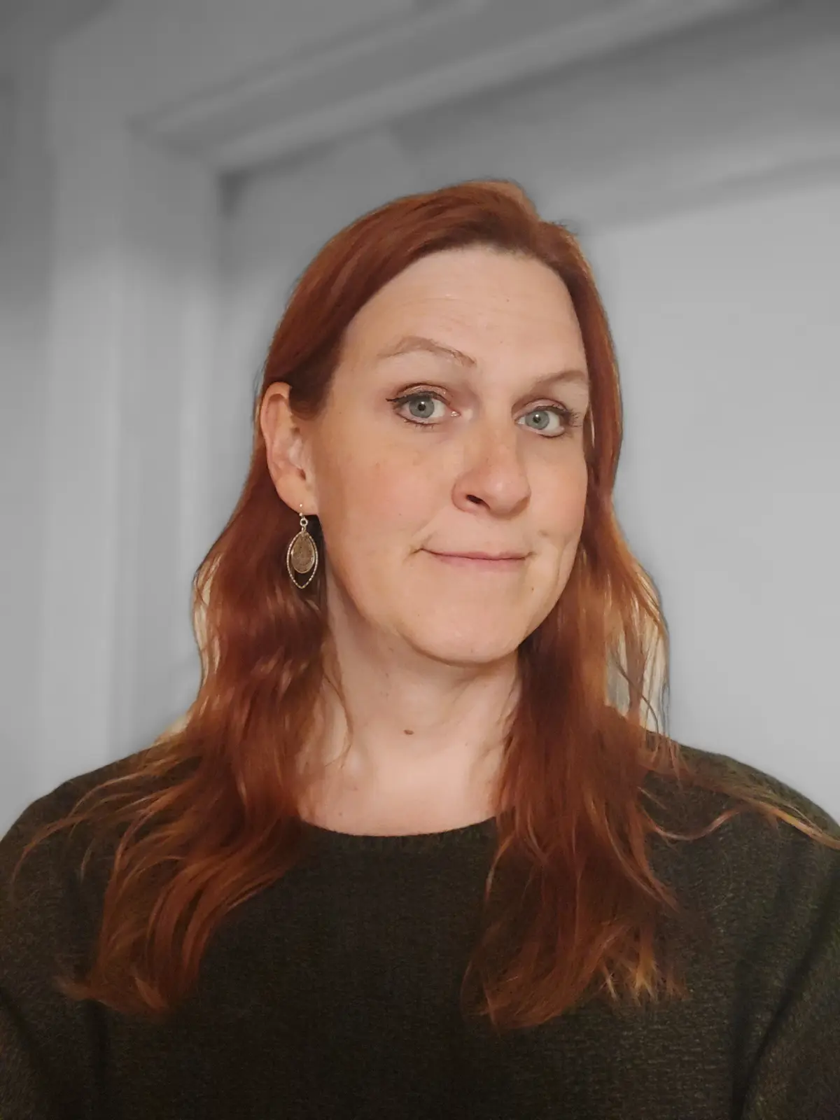 A woman with red hair and a subtle smirk looks at the camera, wearing a dark sweater and dangling earrings, with the text…
