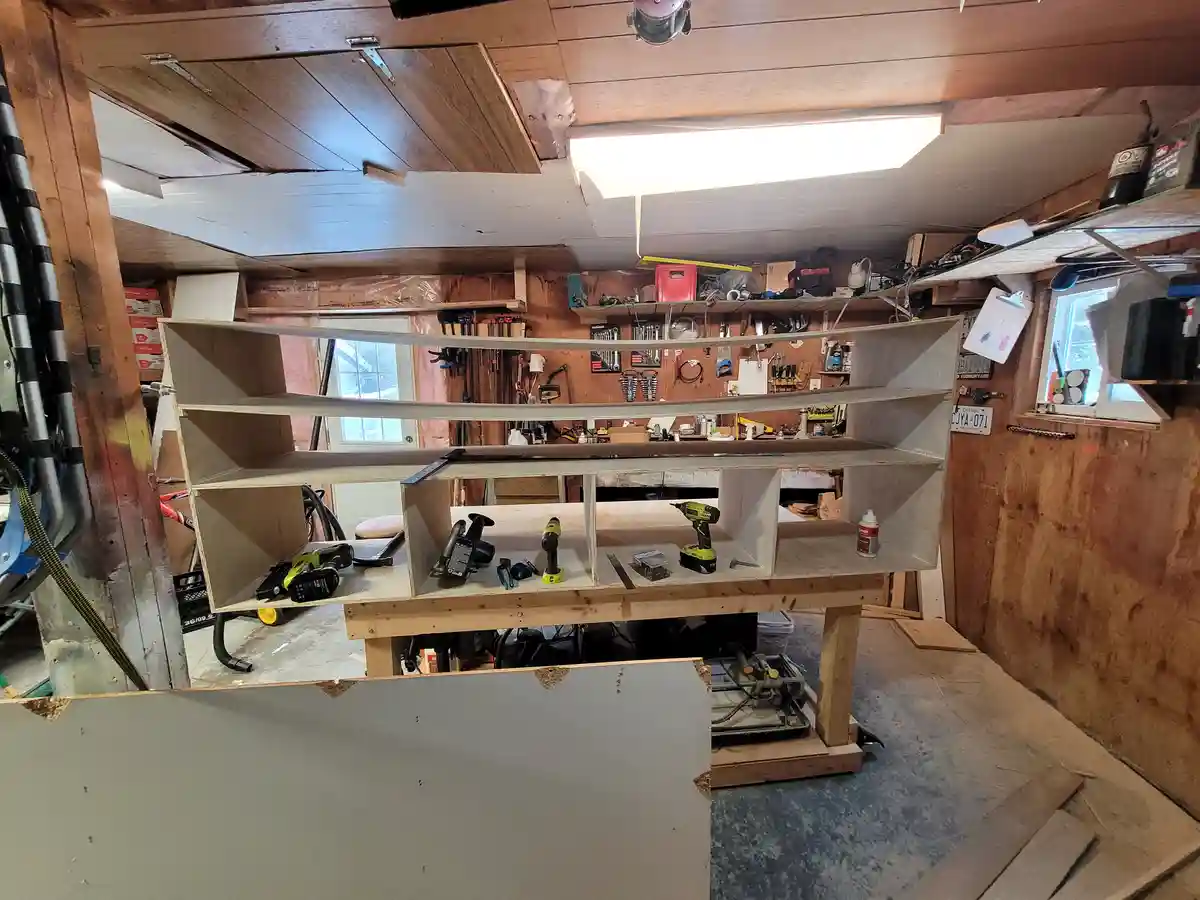 A photo of a cluttered workshop with a large, unfinished wooden shelf unit standing on sawhorses. The shelf has multiple…