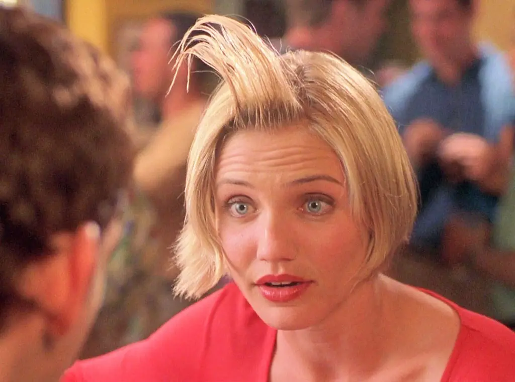 a woman with short blonde hair and a red top stares in shock, her eyebrows furrowed and mouth slightly open, as if reacting…
