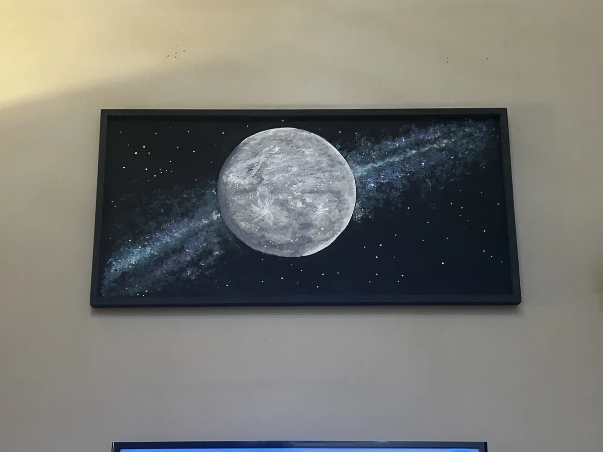 A framed painting of a large, textured moon against a dark, starry sky with a blue nebula swirl hangs on a beige wall. The…