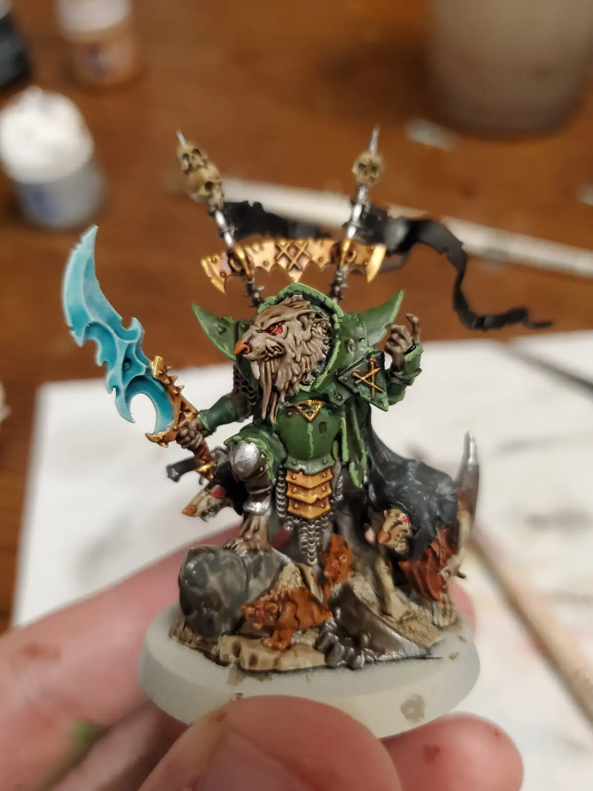 A hand holds a highly detailed, painted miniature of a fantasy warrior with a wolf-like face, green armor, and a blue curved…