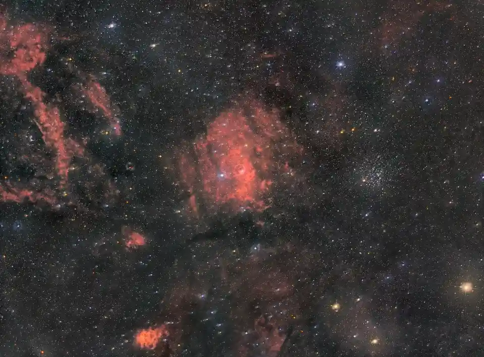 A deep-space photograph shows a dark cosmic field filled with countless stars and glowing red nebulae. Overlaid text reads…