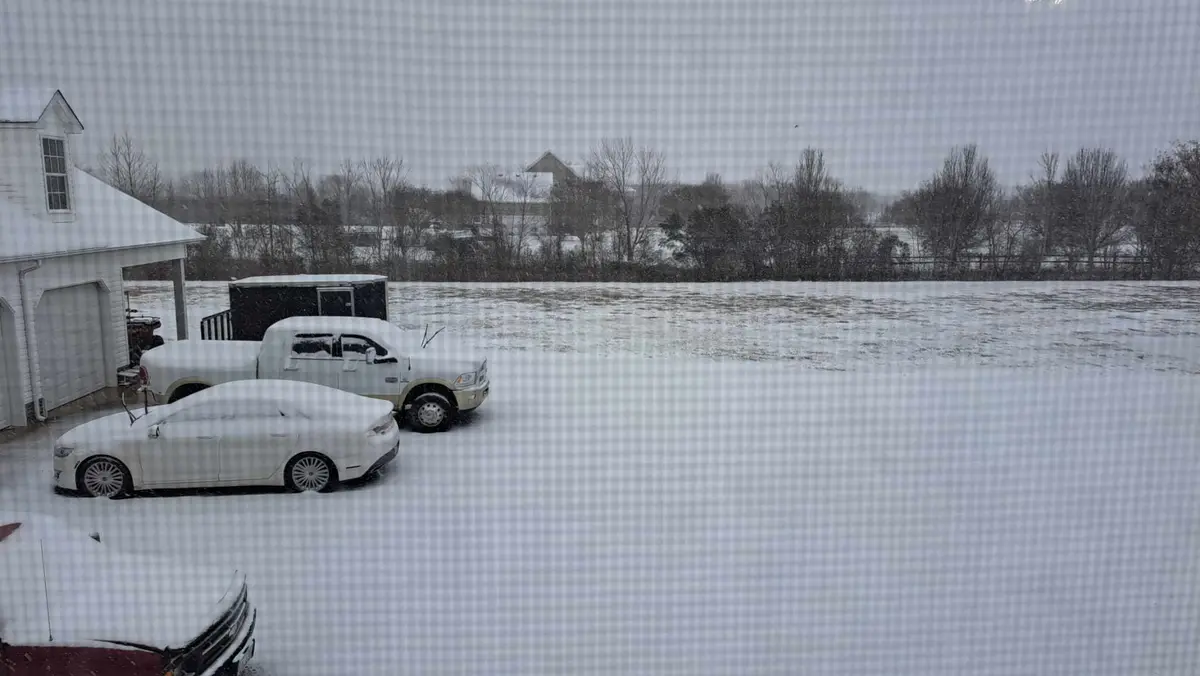 A snowy outdoor scene viewed through a window screen, showing a white car and a white pickup truck parked in front of a…