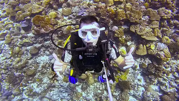A scuba diver in full gear, including a mask, snorkel, and wetsuit, gives two thumbs up while submerged among coral…