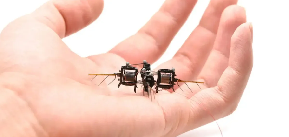 a human hand holds a very small, intricate robotic device resembling a mosquito, with visible wires, motors, and thin legs…