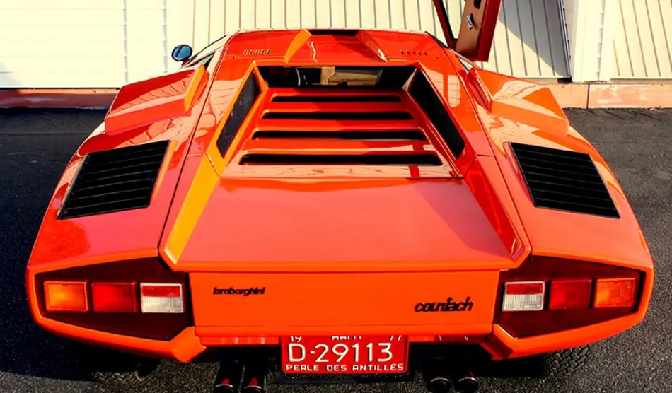 The rear view of a bright orange Lamborghini Countach with its iconic scissor doors open, showing the car's aggressive…