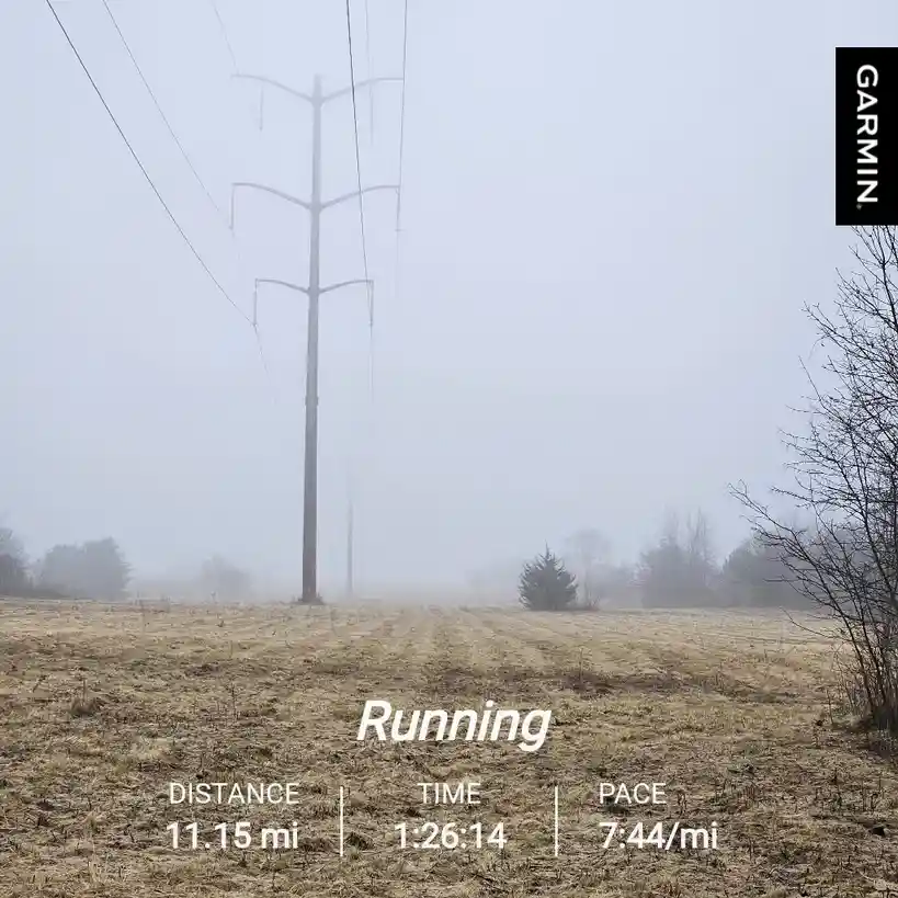 A foggy, desolate field with a tall power line pole in the center, overlaid with a Garmin running stats graphic showing…