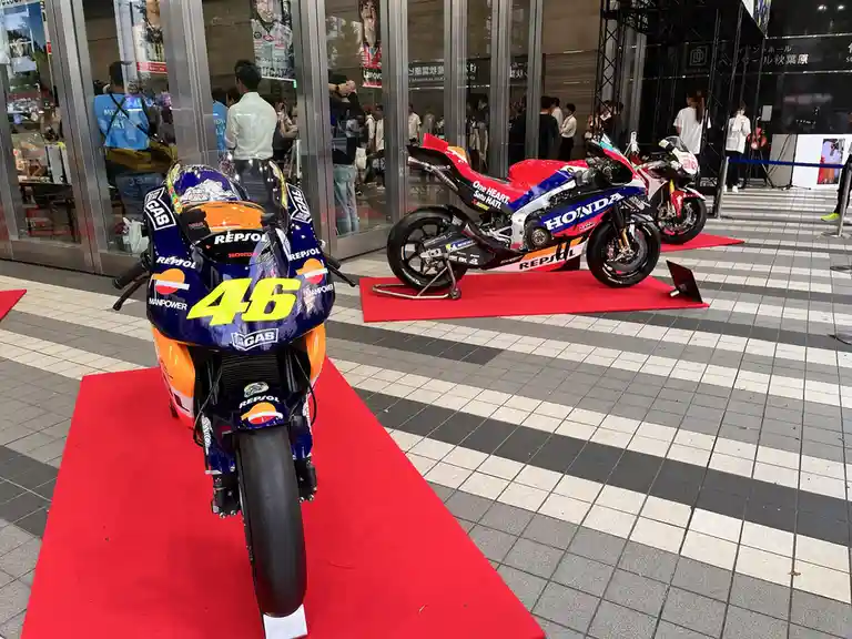 Two racing motorcycles are displayed on red carpets outside a building, one prominently featuring the number 46 and Repsol…