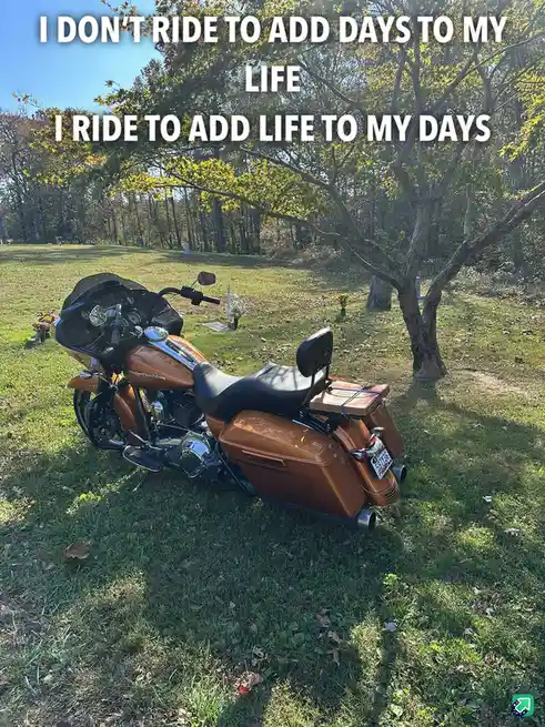 A shiny orange motorcycle is parked on grass in a cemetery, surrounded by trees and headstones under a clear blue sky. The…