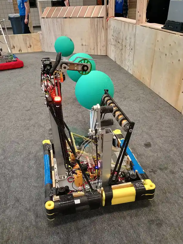 A complex, homemade robot with exposed wiring, gears, and a red light is positioned on a carpeted floor, facing three large…