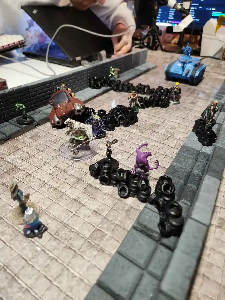 A tabletop wargaming scene shows numerous painted miniatures, including a blue tank, a wrecked red car, and various…