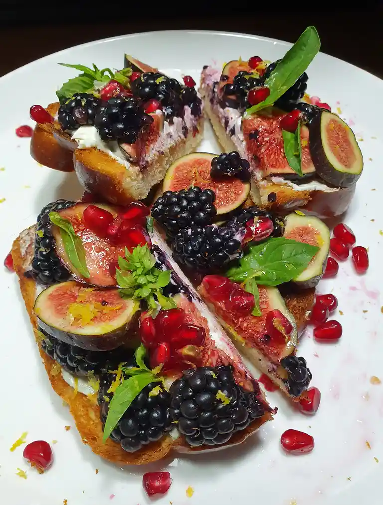 A white plate holds several slices of toasted bread topped with creamy spread, sliced figs, blackberries, pomegranate seeds…
