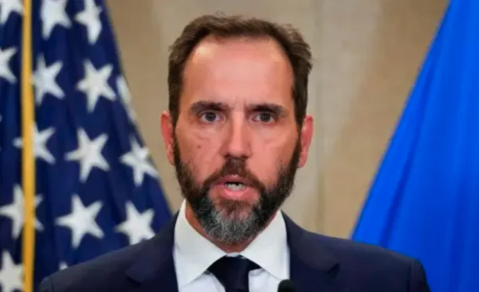 a man with a beard and dark suit stands in front of an american flag and a blue flag, looking shocked or concerned. The…
