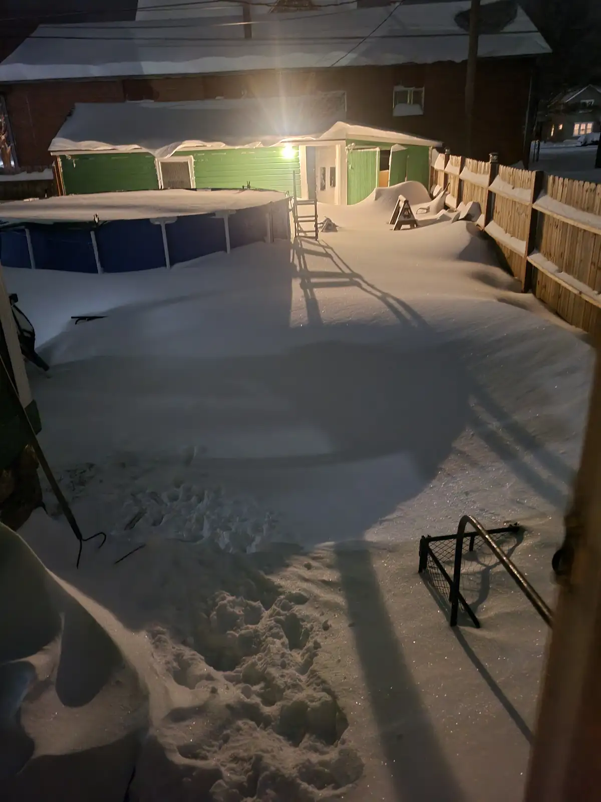A snowy backyard at night illuminated by a bright light, showing a large above-ground pool completely covered in snow, a…