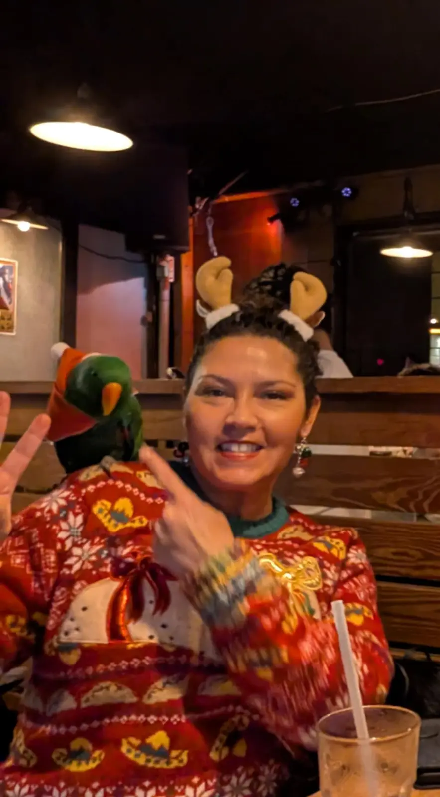 A woman wearing a festive red sweater with taco and snowflake patterns and reindeer antlers sits in a dimly lit restaurant…