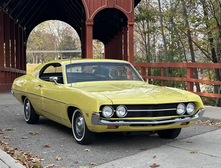 A bright yellow classic Ford muscle car, likely a 1969-1970 model, is parked under a red wooden covered bridge. The car has…
