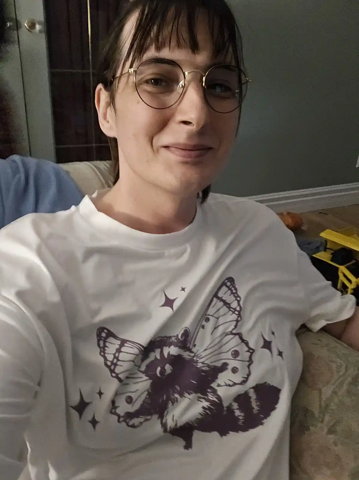 A person with short dark hair and round glasses smiles while wearing a white t-shirt featuring a purple graphic of a raccoon…