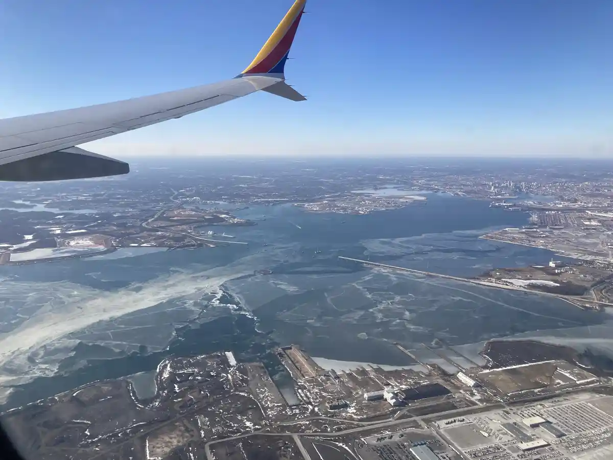 An aerial view from an airplane window shows the wing with a red, yellow, and blue tail fin against a clear blue sky. Below…