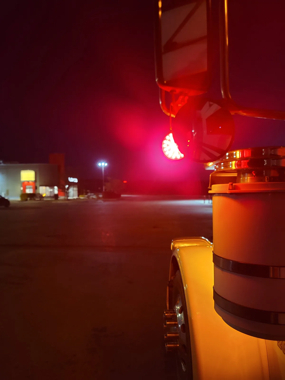 A nighttime photo from the side of a large truck, showing its red cab lights glowing brightly. The truck's side mirror…