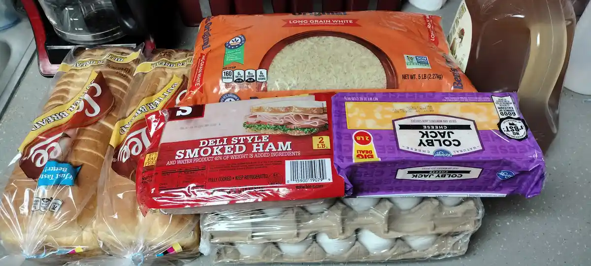A kitchen counter displays groceries including butter bread, a large bag of long grain white rice, a package of deli style…