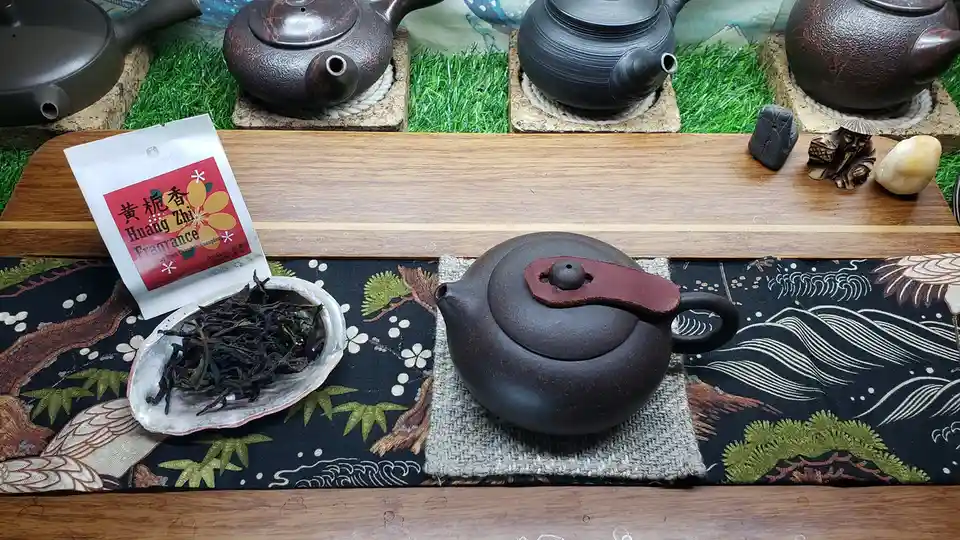 A still life of a dark brown ceramic teapot with a leather handle sits on a woven mat atop a patterned table runner. Beside…
