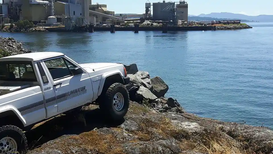 A white, lifted Jeep Cherokee Commander is parked precariously on a rocky shoreline next to a body of water, with industrial…