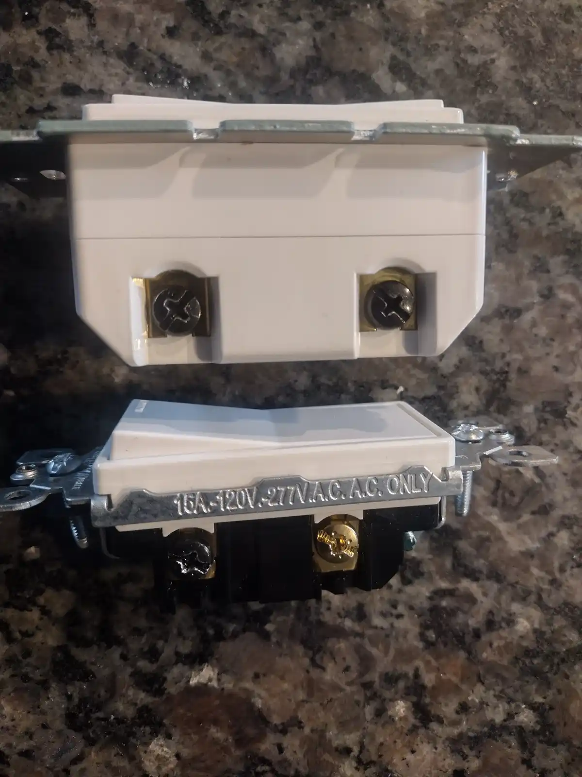 Two electrical outlets are shown back-to-back on a granite countertop, one white and one black, both with visible screw…