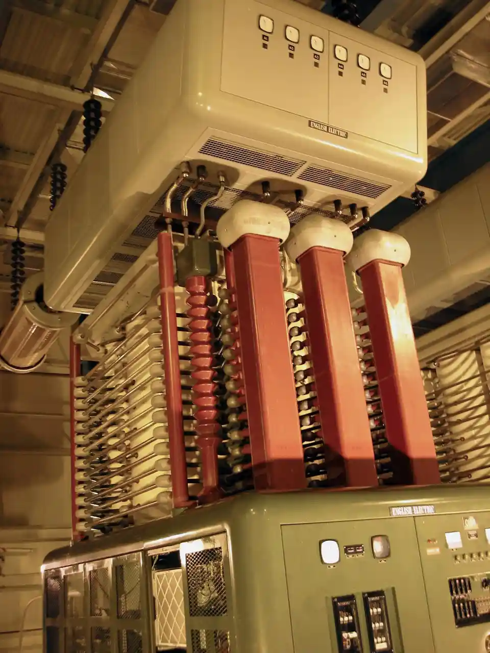 A low-angle photo of vintage English Electric electrical equipment, featuring large red insulators and control panels…