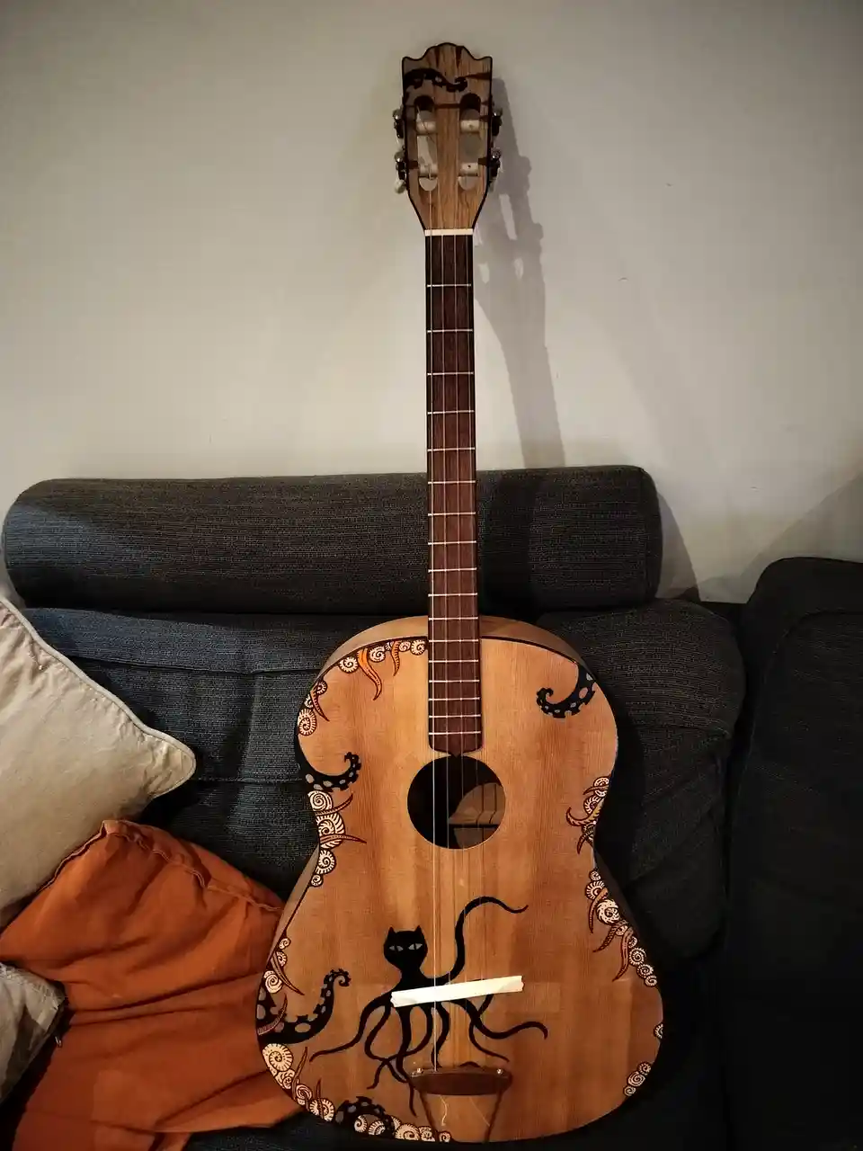 A wooden acoustic guitar with a black cat-octopus design painted on its body, leaning against a dark gray couch with…