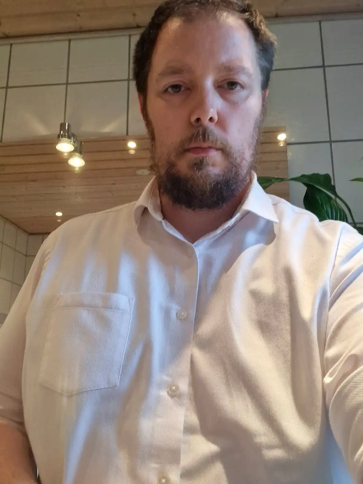 A man with a beard and short hair, wearing a white button-up shirt, stares directly at the camera with a neutral expression…