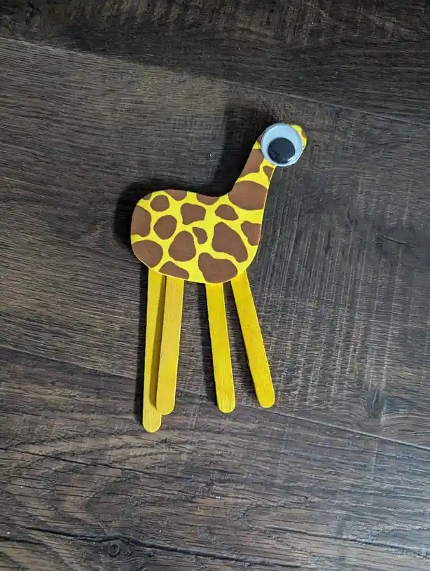 A handmade giraffe craft made from a yellow and brown spotted paper cutout body, a single large googly eye, and four…