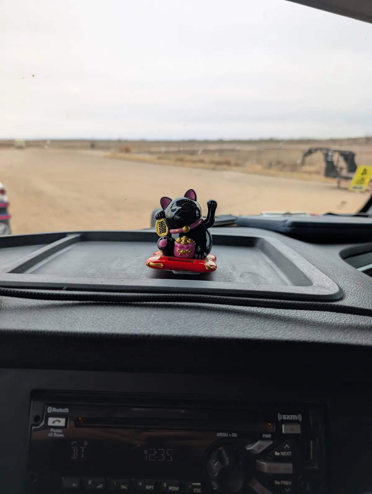 A black and pink lucky cat figurine sits on a car dashboard, one paw raised in a traditional 'beckoning' gesture, while the…