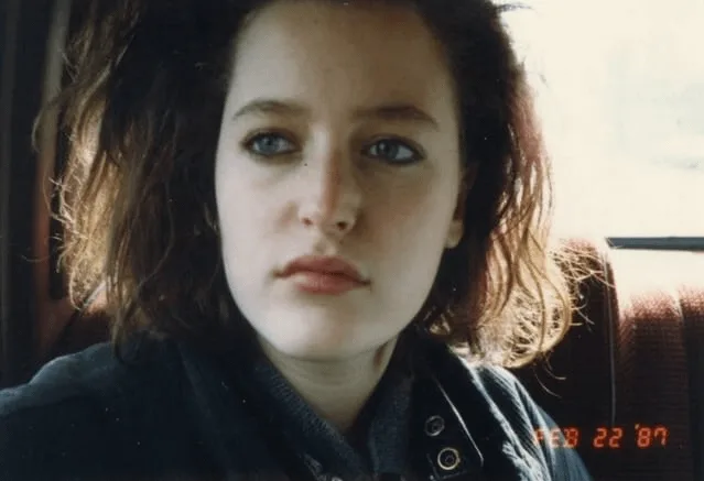 A close-up photo of a young woman with wavy brown hair and blue eyes, looking directly at the camera with a serious…