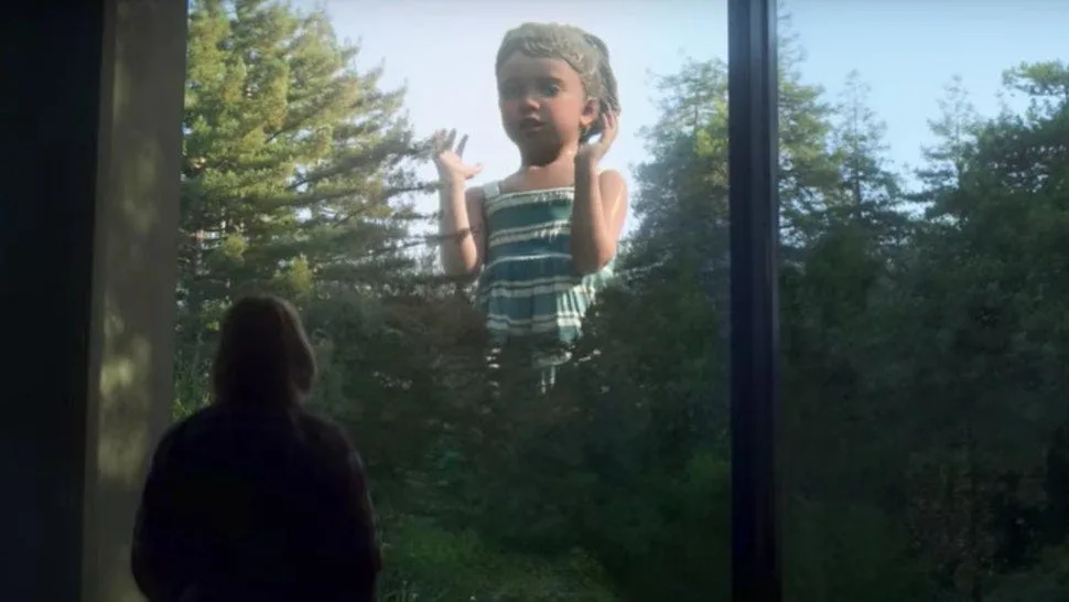 A person viewed from behind stands inside a room, looking out a large window at a giant, distorted image of a young girl in…