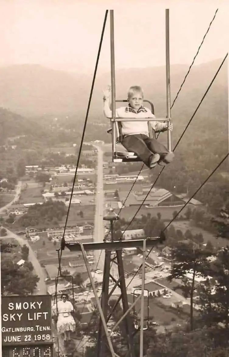 A vintage black-and-white photo from June 22, 1958, shows a young boy sitting in a chairlift suspended high above…