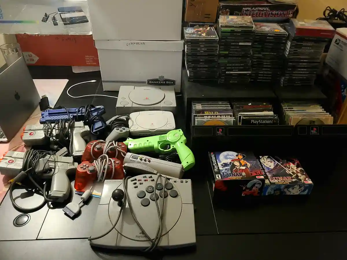 A cluttered desk displays a large collection of PlayStation 1 hardware and games, including multiple consoles, controllers…