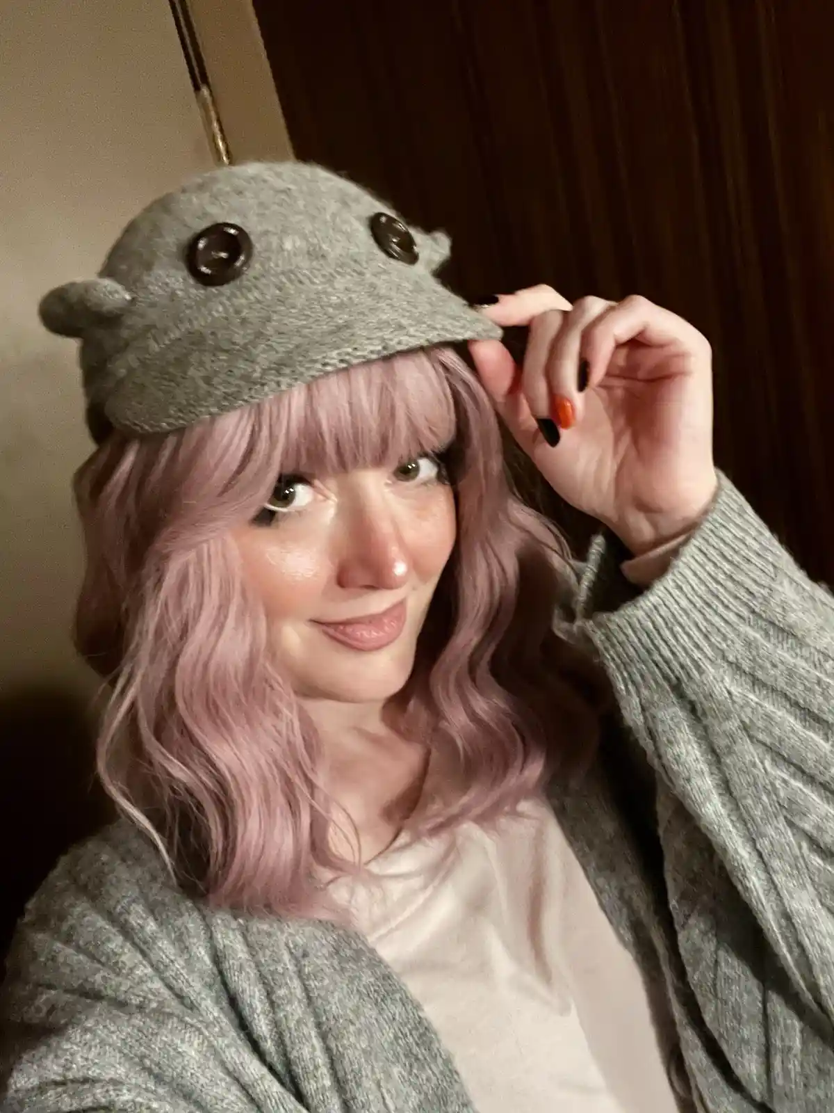 A person with pink hair and a grey knitted cardigan wears a grey felt hat shaped like a hippo, complete with button eyes and…