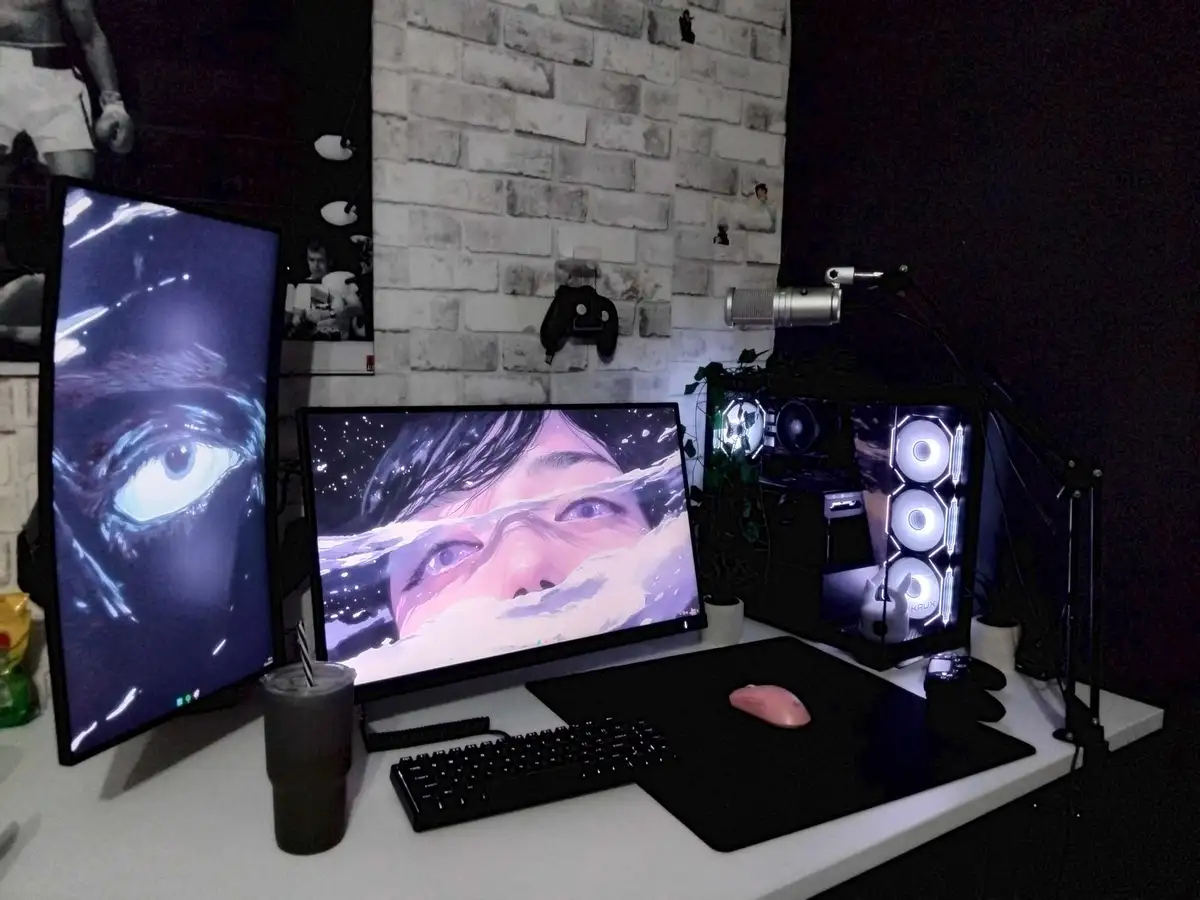 A dimly lit gaming setup with two monitors displaying artistic eye wallpapers, a glowing custom PC case, a pink mouse, and a…