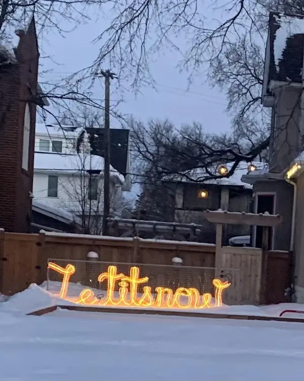 A snowy backyard scene features a glowing orange neon sign spelling 'Let it snow' lying in the snow on the ground. Bare…
