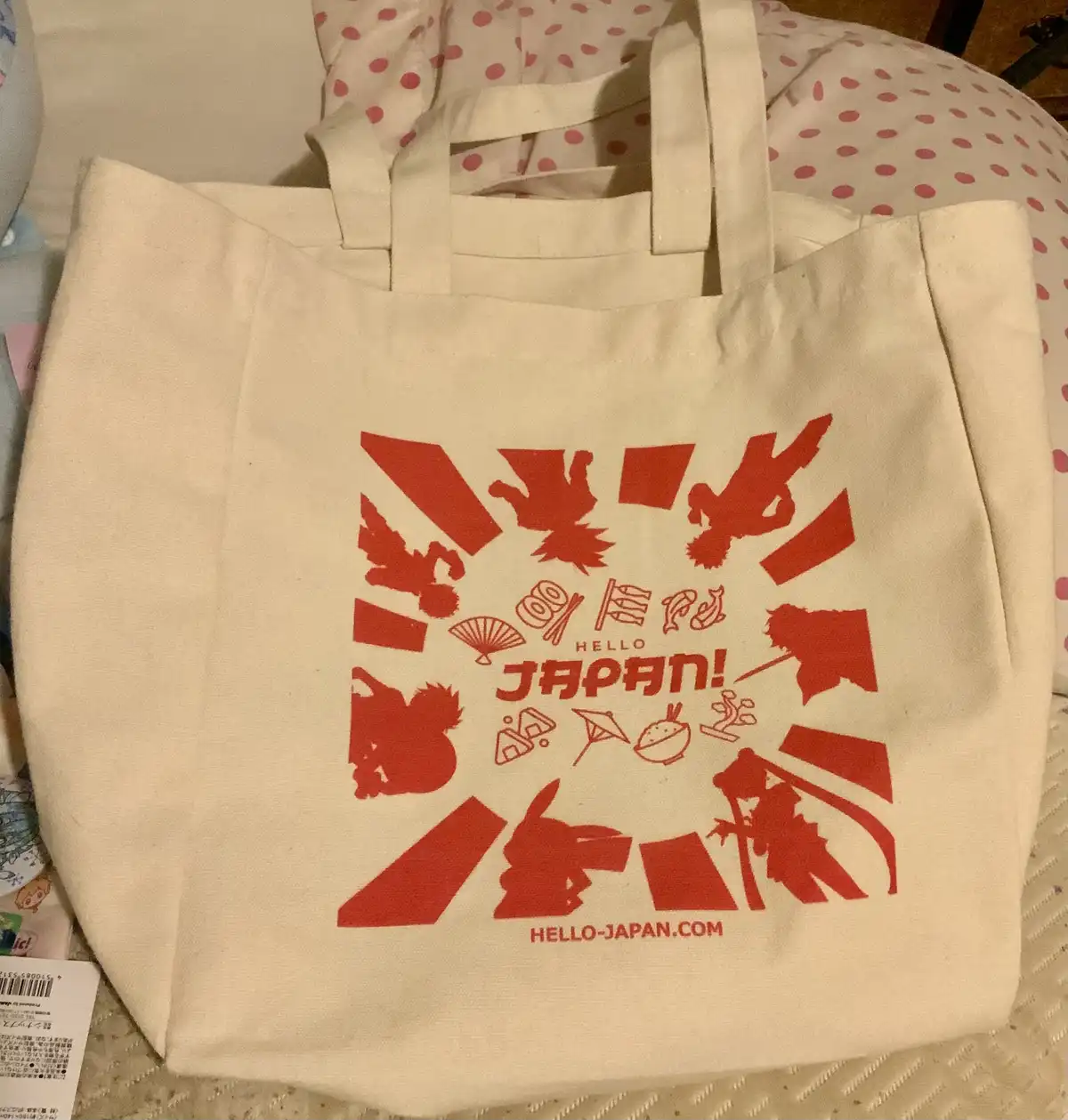 A beige tote bag with a red 'Hello Japan!' logo and various Japanese cultural icons like fans, kites, and food, sits on a…